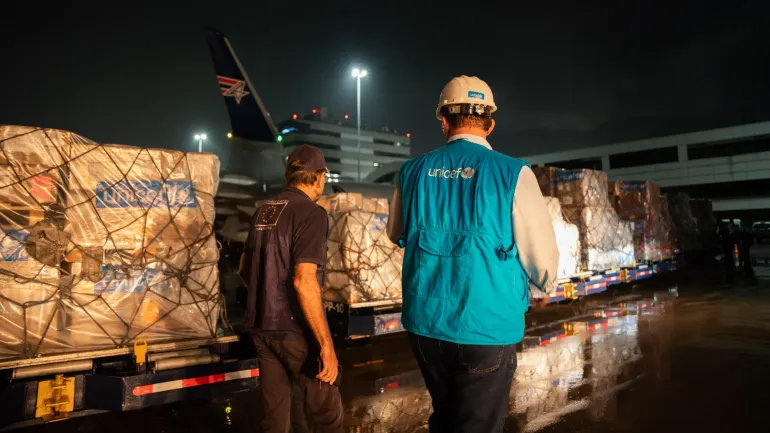 An EU humanitarian worker and a UNICEF staff member walk past a shipment of humanitarian aid supplies at night.