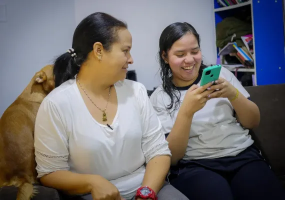 Una mujer y una adolescente sentadas en un sofa, la mujer adulta mira a la adolescente mientras ella usa un tel&eacute;fono celular.