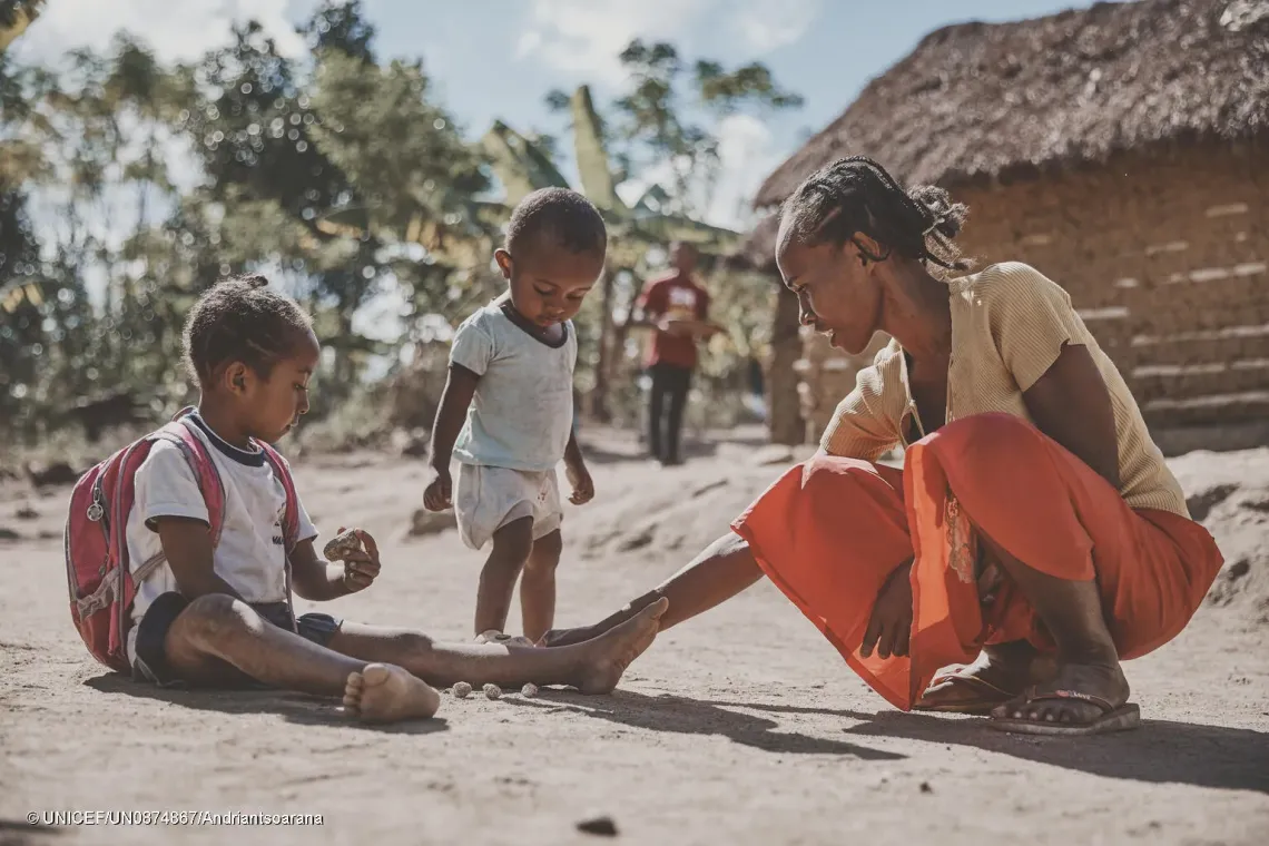 Jos&eacute;phine Kezia, parent-leader, joue avec sa fille devant leur maison, tandis que leur plus jeune enfant se tient &agrave; leurs c&ocirc;t&eacute;s. &Agrave; l'arri&egrave;re-plan, on aper&ccedil;oit son mari pr&egrave;s de la maison familiale.