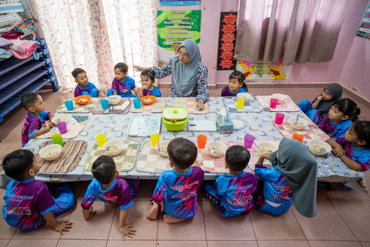 Children sit together with their teacher, Cikgu Maizon, to enjoy a nutritious breakfast prepared at their childcare centre in Gua Musang, Kelantan. Shared mealtimes support healthy eating habits, routine and social learning during early childhood.