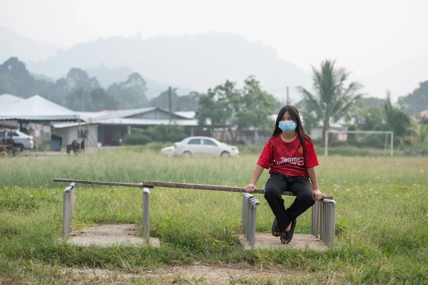Eva sits alone on a simple bench in an open field, wearing a face mask as a thick haze softens the landscape behind her. Homes, trees and hills fade into the hazy sky, showing how air pollution has become part of the environment around where she lives.