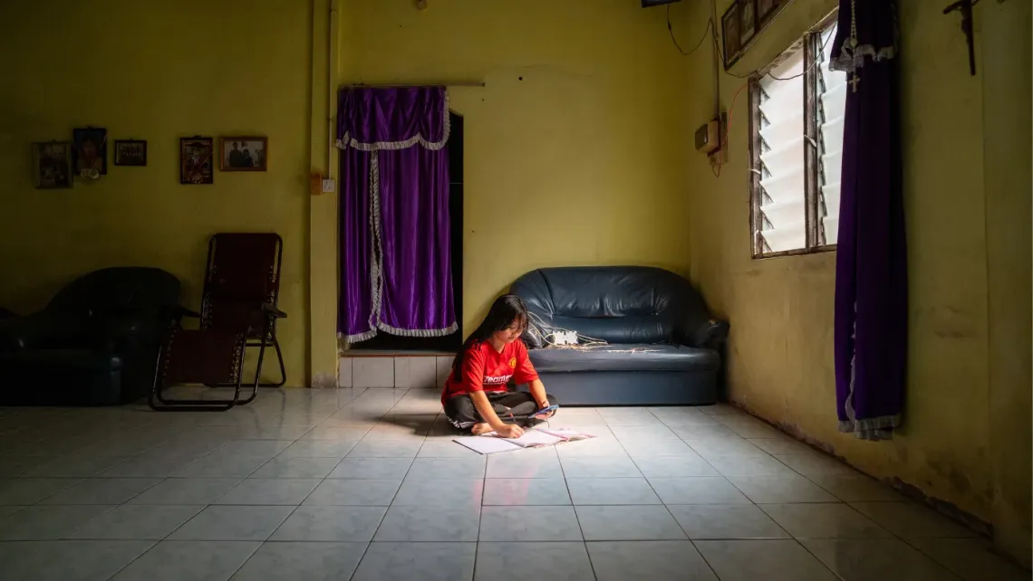 Eva studies on the floor of her home, using her phone and schoolbooks to continue learning during a school closure caused by severe haze.