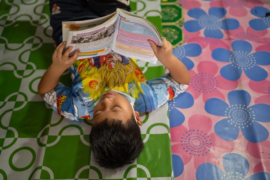 Andrea lies on the floor at home reading a schoolbook, with his face mask lowered beneath his chin.