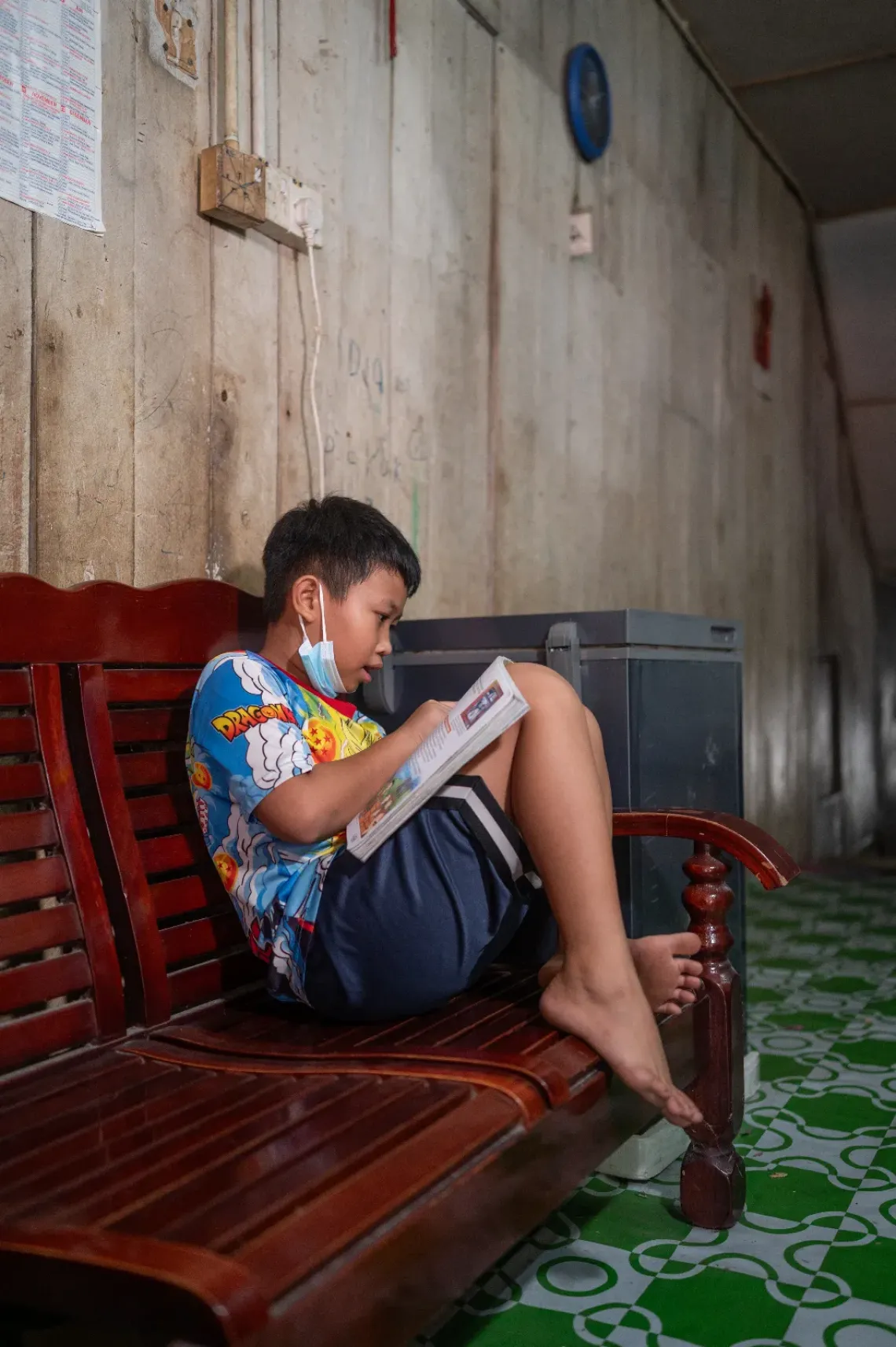 Andrea sits on a wooden bench at home, reading a book with a face mask lowered beneath his chin.