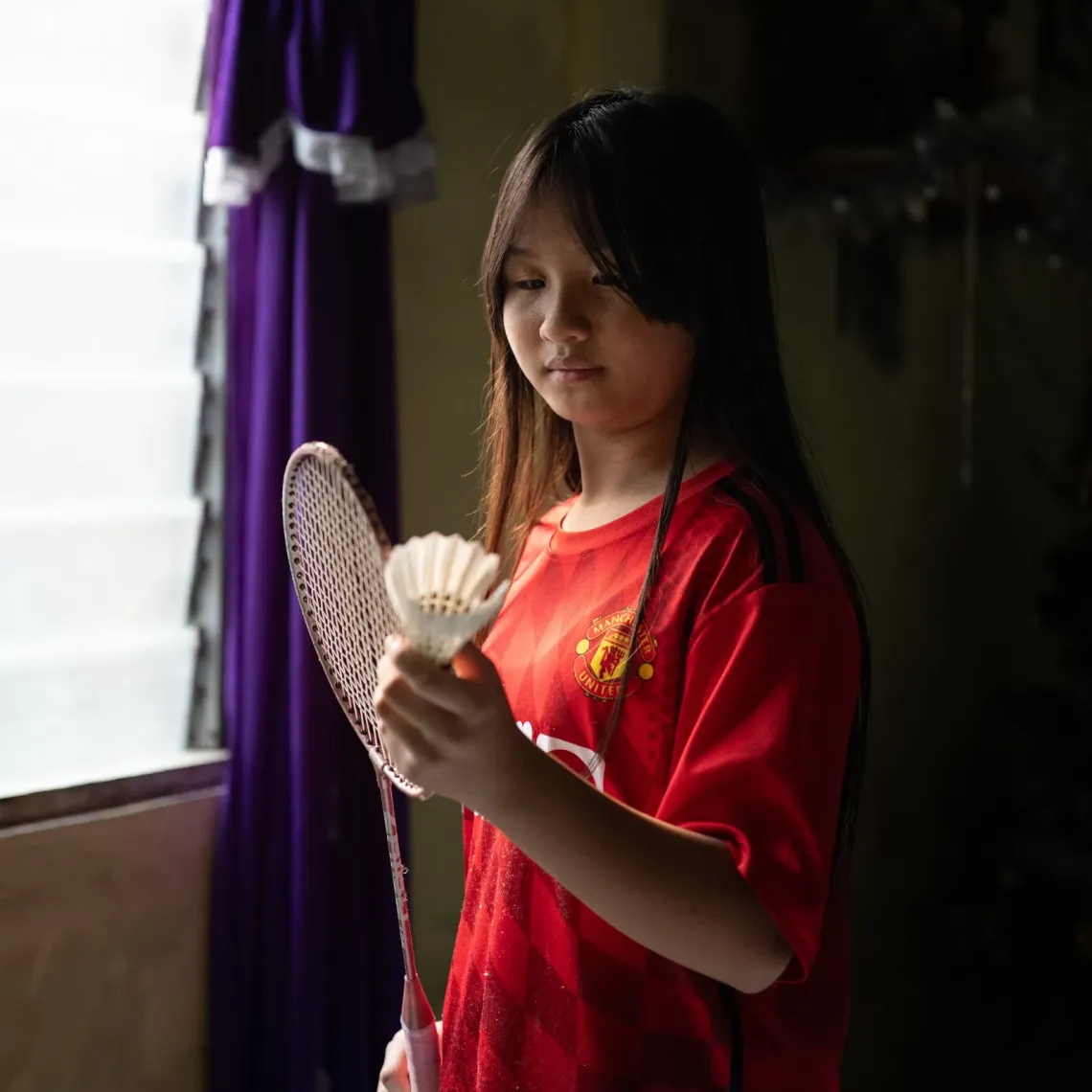 Eva holds a badminton racket and shuttlecock while standing by a window at her home in Kampung Mongkos, Serian, Sarawak.