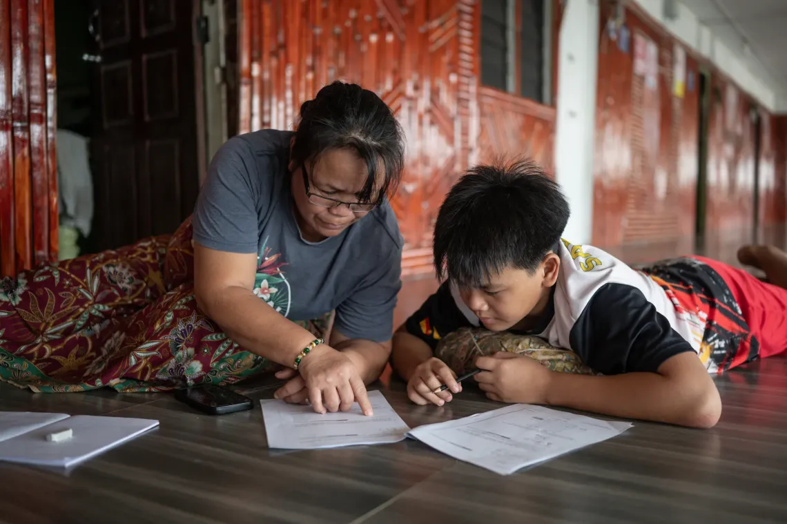 Freddy lies on the floor beside his mother as she helps him with his schoolwork at home.