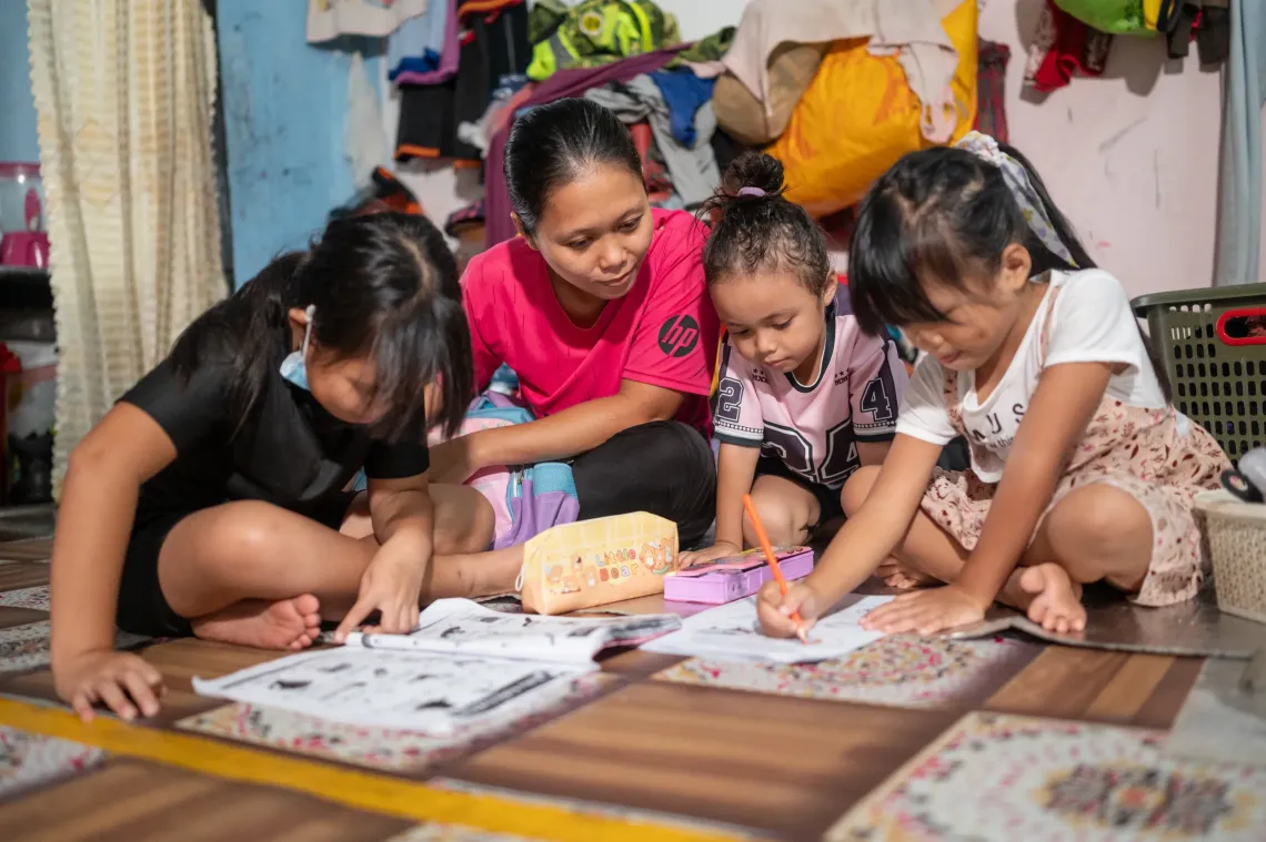 Zara sits on the floor with her mother and two younger children as they study and complete schoolwork together at home.