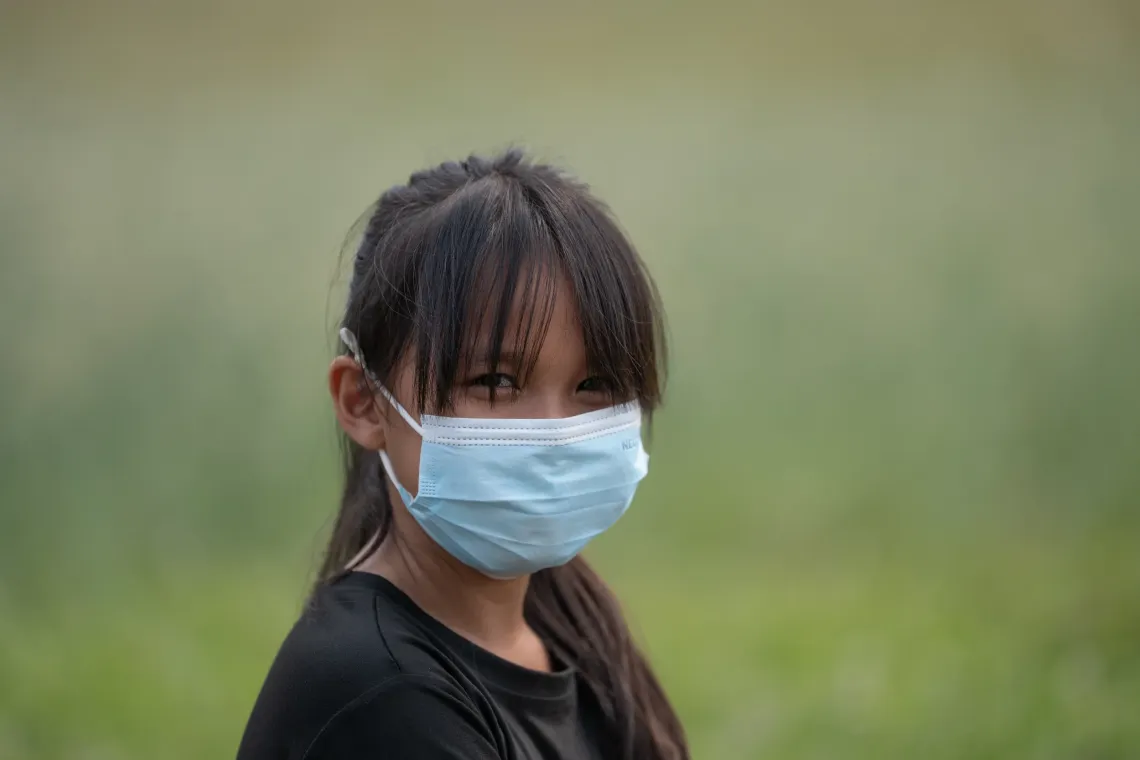 A young girl wearing a face mask, stands outdoors against a blurred green background.