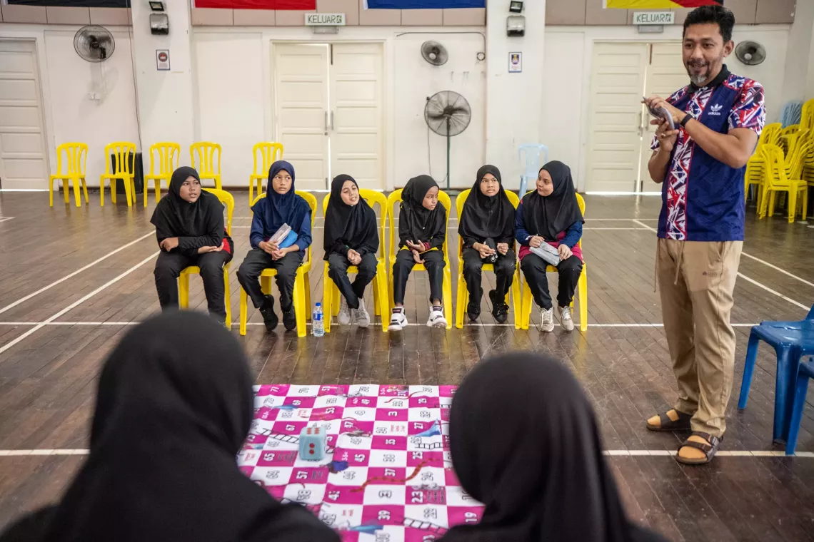 6 teen girls sit in the middle of a gym awaiting instructions from their teacher.