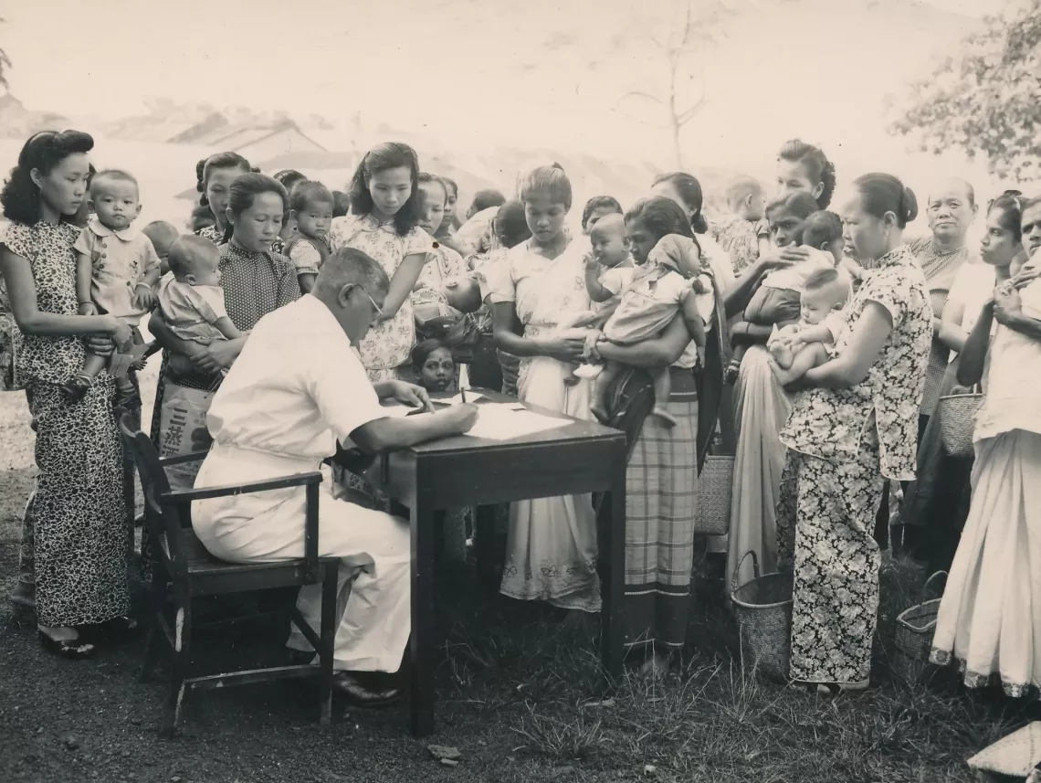 A group of mothers and babies surround a clerk seated at a table. 