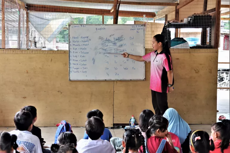 A young teacher points to words on a white board while her students are seated on the floor. 