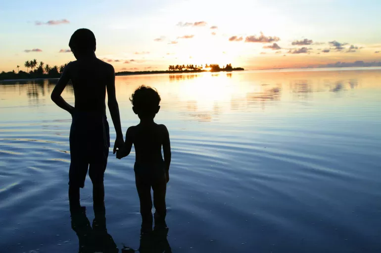 A young boy and his brother stand in front of the ocean at sunset.