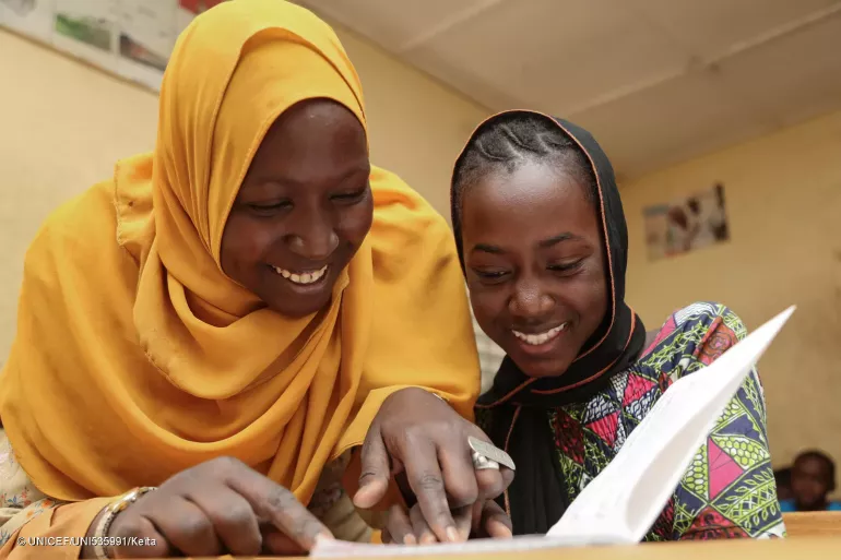 A teacher stands next to a child that is sitting at a school desk. The child is reading a book. Both are smiling. 