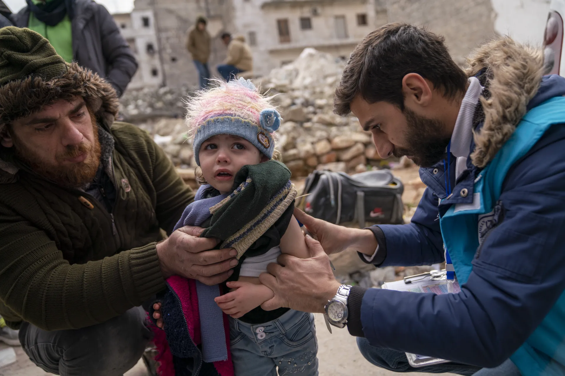 Male health team worker looks at a toddler's arm with rubble from a collapsed building in the background.