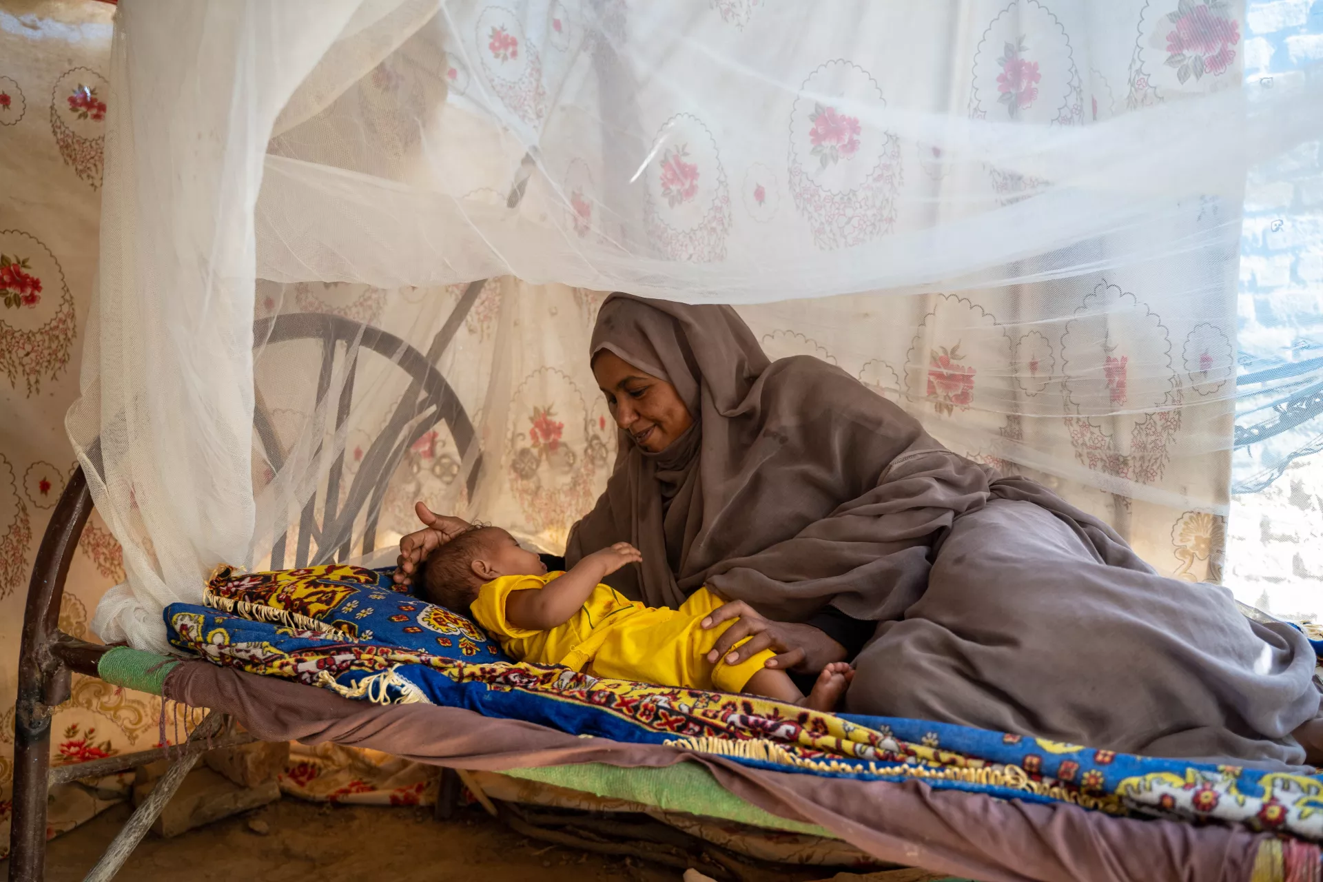 On 22 February 2023, mother Sabna Mohamed lies in bed beside her 9-month-old daughter, Fatuma, in Wager village, North Delta locality, Kassala State, Sudan, as Fatuma nears naptime.