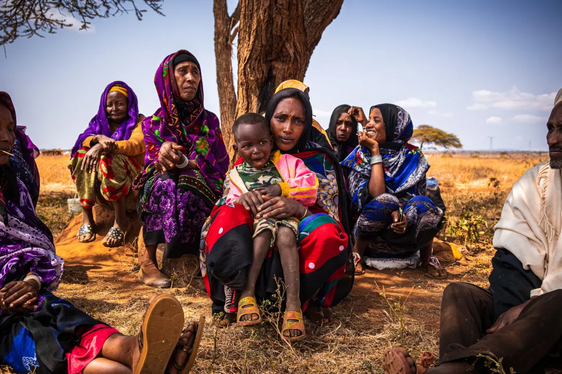 Three-year-old Derara sits on his mother's lap under a tree while surrounded by other community members.