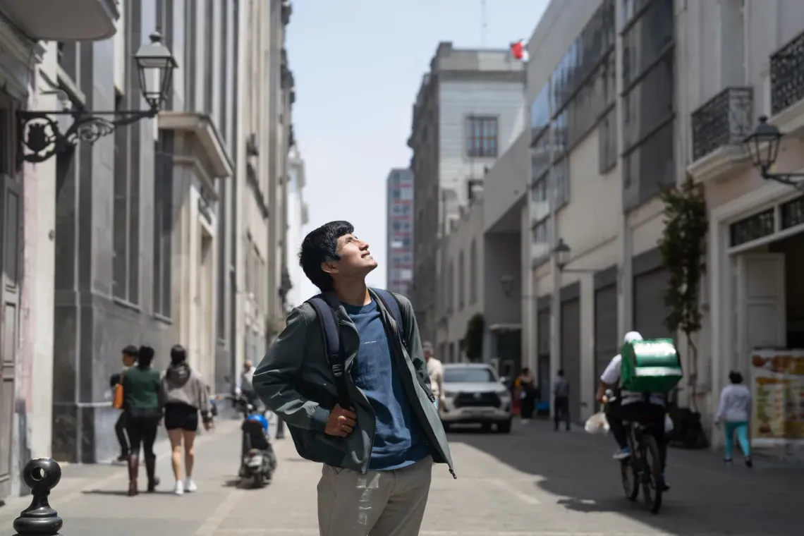 A teenage boy wearing a backpack looks upward while standing on a sunny city block in Lima, Peru.