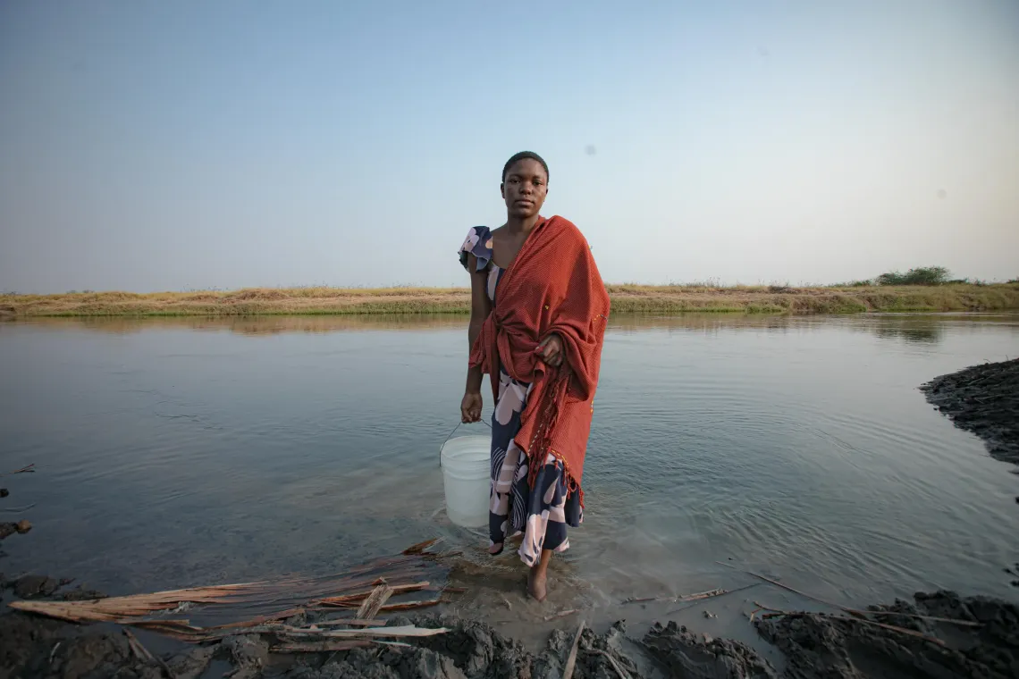 Antini, dressed in a red shawl walks out of a lake carrying a bucket of water. 