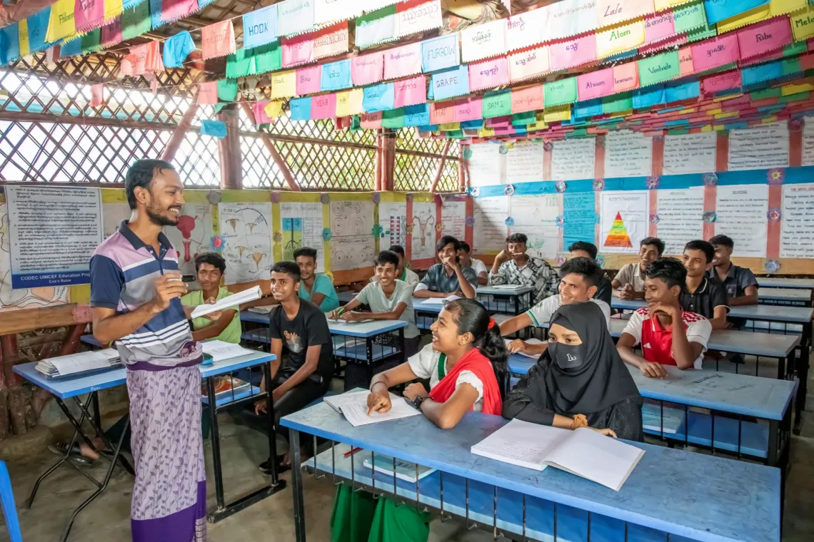 Kulsum sits at a blue desk in a classroom of children, smiling with her teacher.