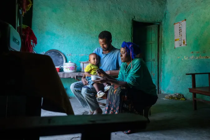 A couple holding and feeding their baby inside their clay-walled home