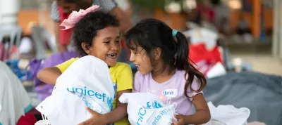  Dos ni&ntilde;as peque&ntilde;as sonriendo y sosteni&eacute;ndose mutuamente mientras cargan bolsas con el logo de UNICEF y art&iacute;culos de higiene
