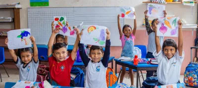 Grupo de ni&ntilde;os y ni&ntilde;as en un sal&oacute;n de clases sosteniendo en alto dibujos de frutas y verduras de colores