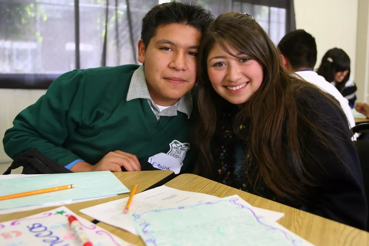 Dos estudiantes j&oacute;venes, un ni&ntilde;o y una ni&ntilde;a, sonr&iacute;en frente a un escritorio que tiene sus apuntes escolares