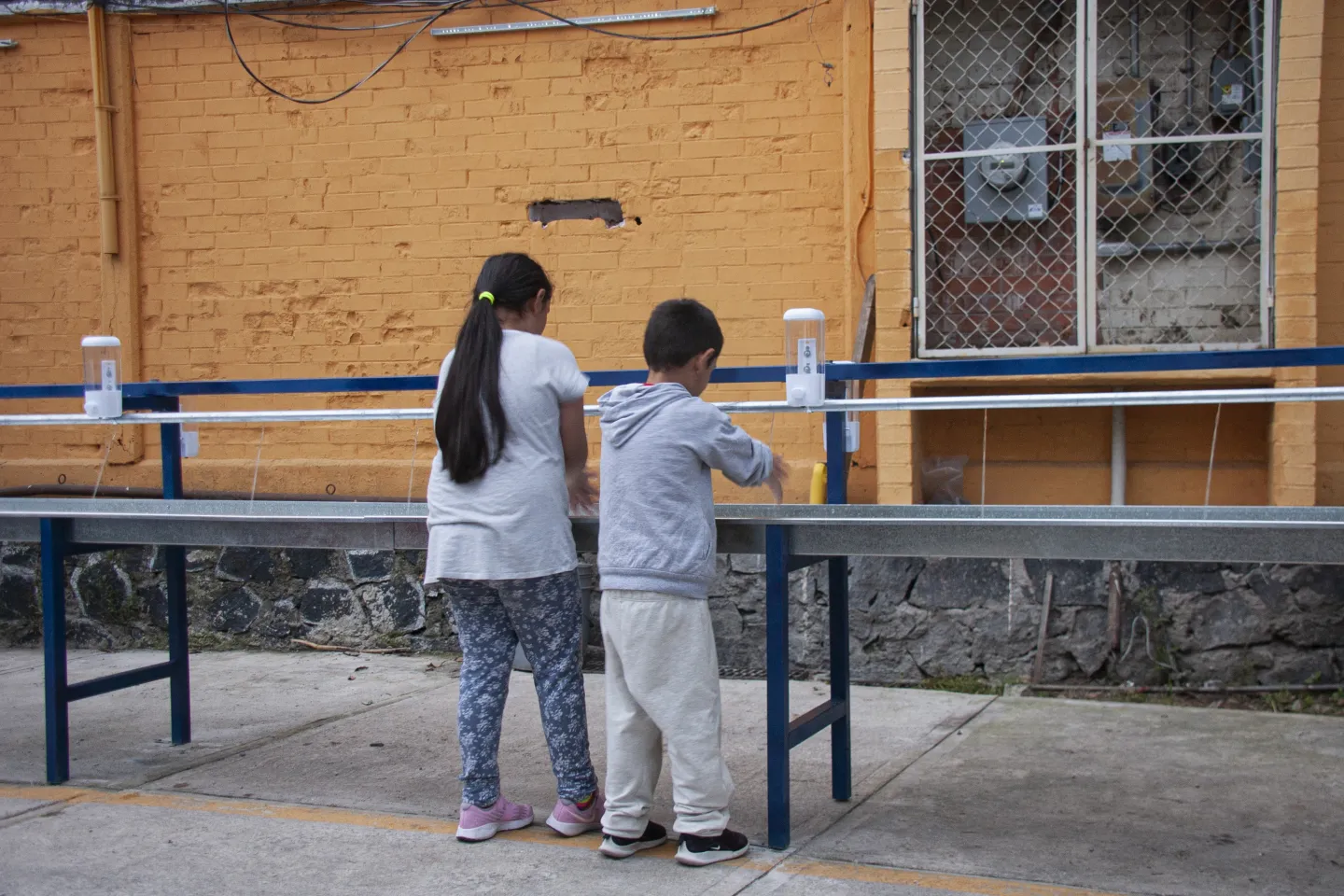 Dos ni&ntilde;os de espaldas lav&aacute;ndose las manos en una estaci&oacute;n de higiene exterior instalada en el patio de una escuela.Dos ni&ntilde;os de espaldas lav&aacute;ndose las manos en una estaci&oacute;n de higiene exterior instalada en el patio de una escuela.