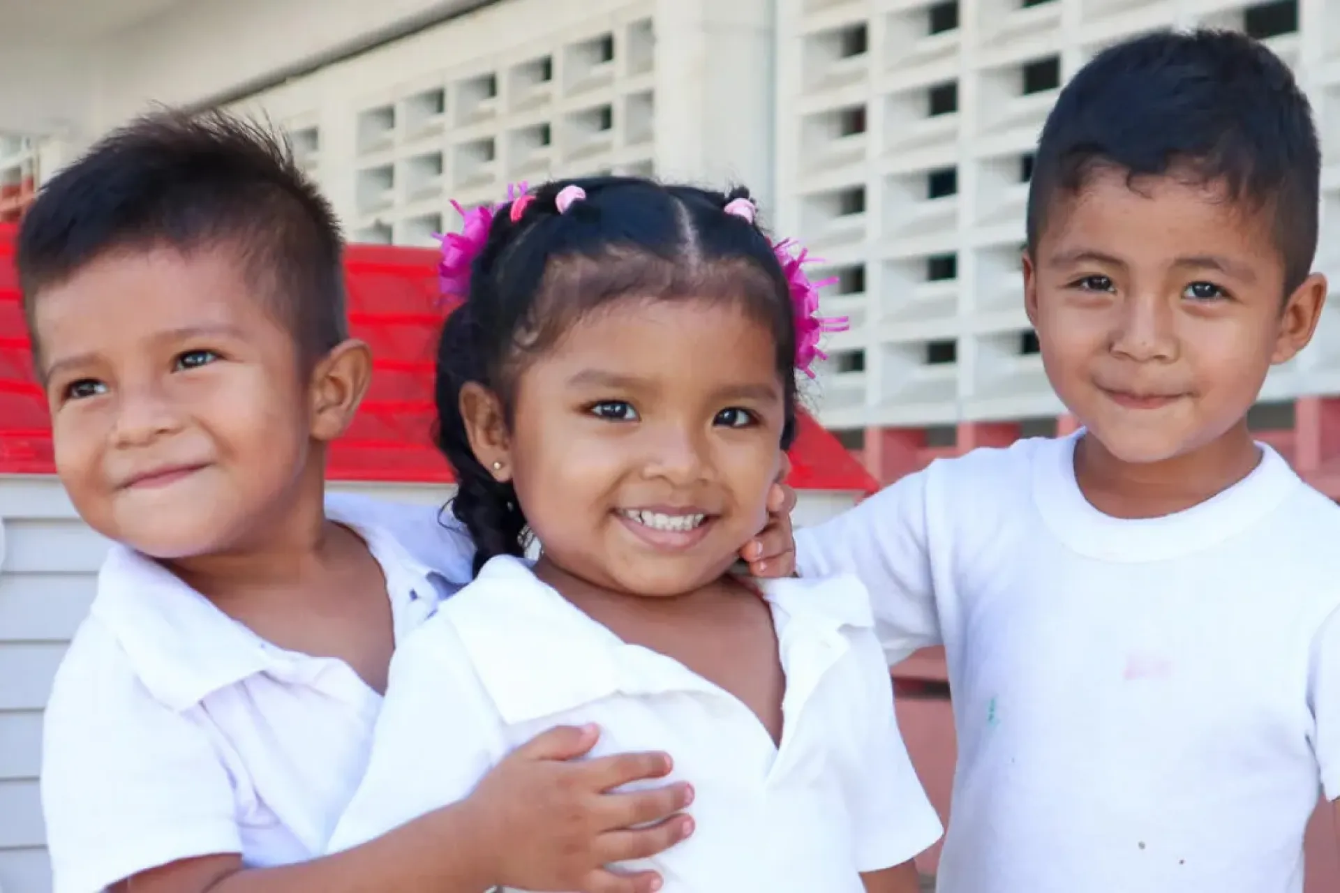 Tres ni&ntilde;os peque&ntilde;os con uniforme escolar blanco sonr&iacute;en abrazados frente a una casita de juguete de pl&aacute;stico en un patio escolar.