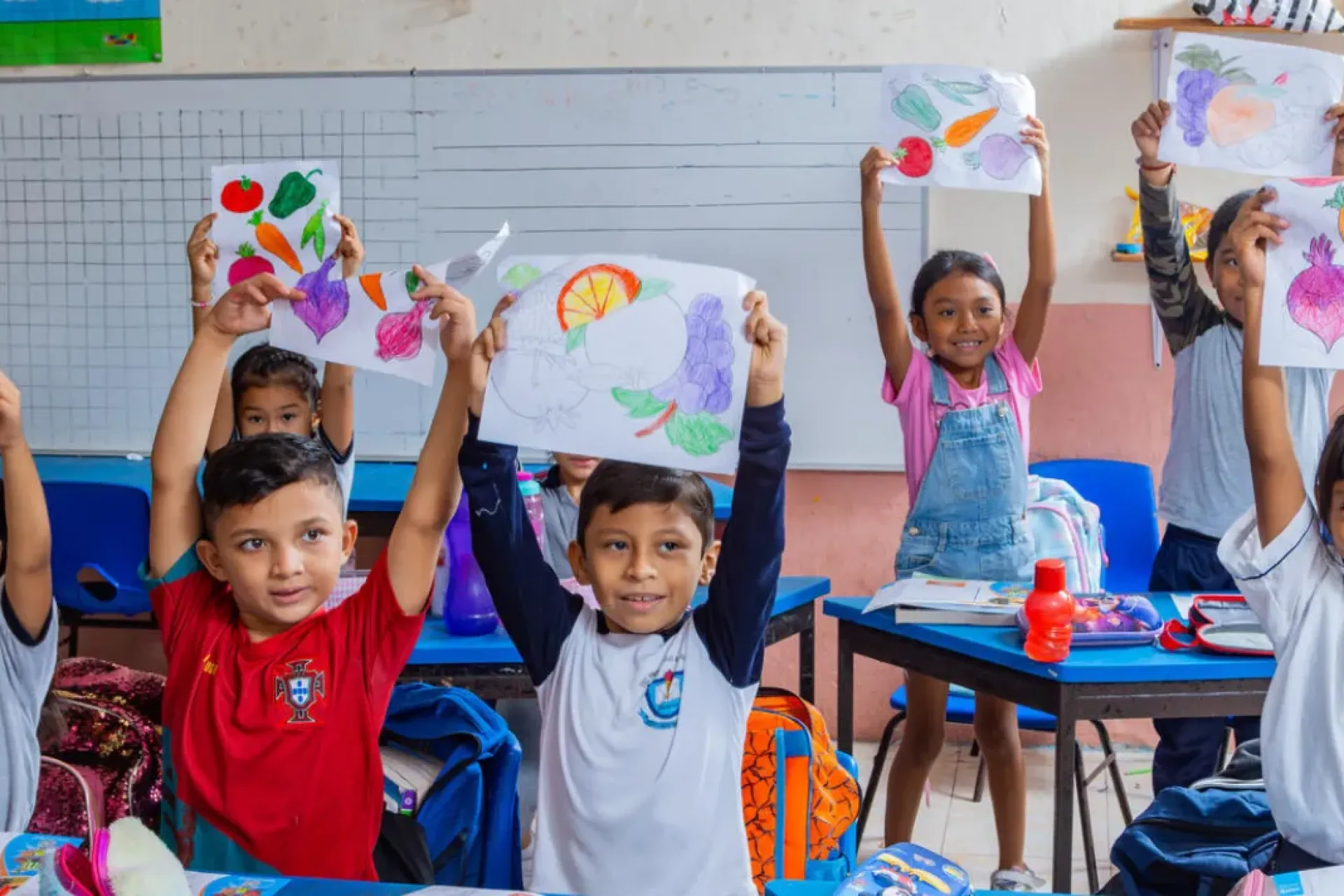 Grupo de ni&ntilde;os y ni&ntilde;as en un sal&oacute;n de clases sosteniendo en alto dibujos de frutas y verduras de colores