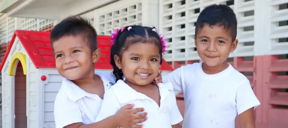 Tres ni&ntilde;os peque&ntilde;os con uniforme escolar blanco sonr&iacute;en abrazados frente a una casita de juguete de pl&aacute;stico en un patio escolar.
