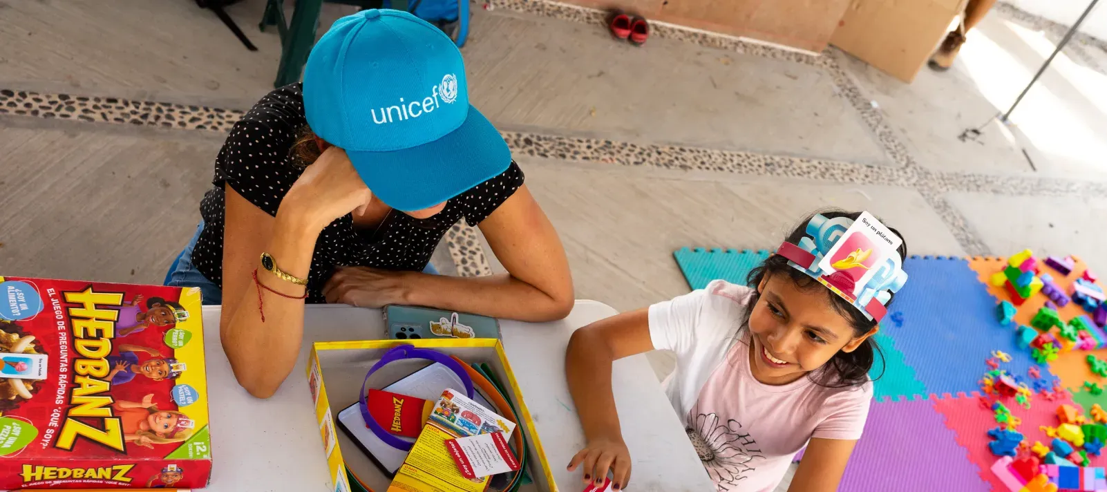 Trabajadora de UNICEF con gorra azul juega con una ni&ntilde;a a un juego de mesa en un espacio amigable con la ni&ntilde;ez..