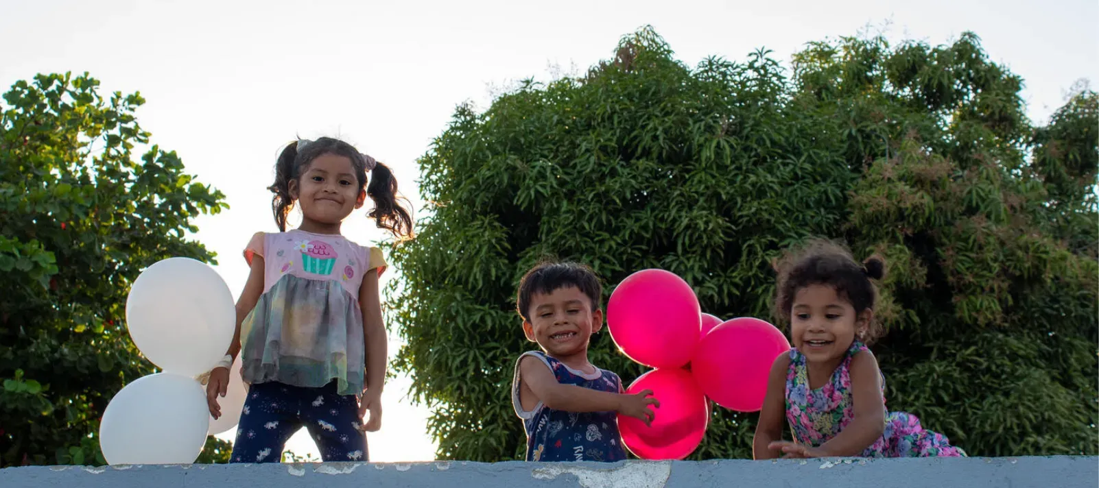 Tres ni&ntilde;os peque&ntilde;os sonr&iacute;en sosteniendo globos blancos y rosas al aire libre.