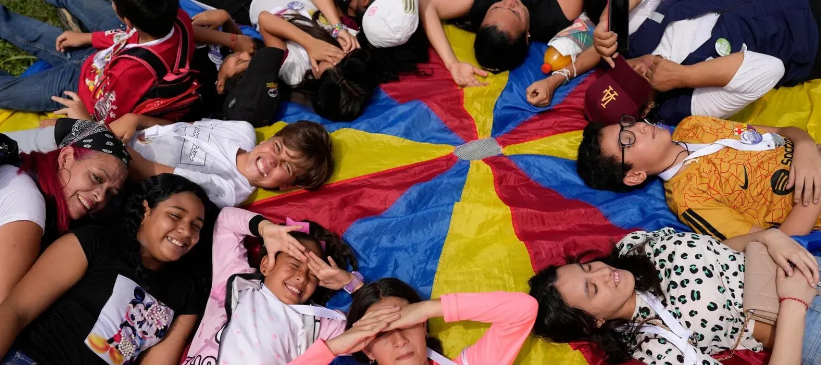 Grupo de ni&ntilde;as, ni&ntilde;os, adolescentes y personas adultas recostados en c&iacute;rculo sobre una tela de paraca&iacute;das de colores, sonriendo y jugando juntos al aire libre.