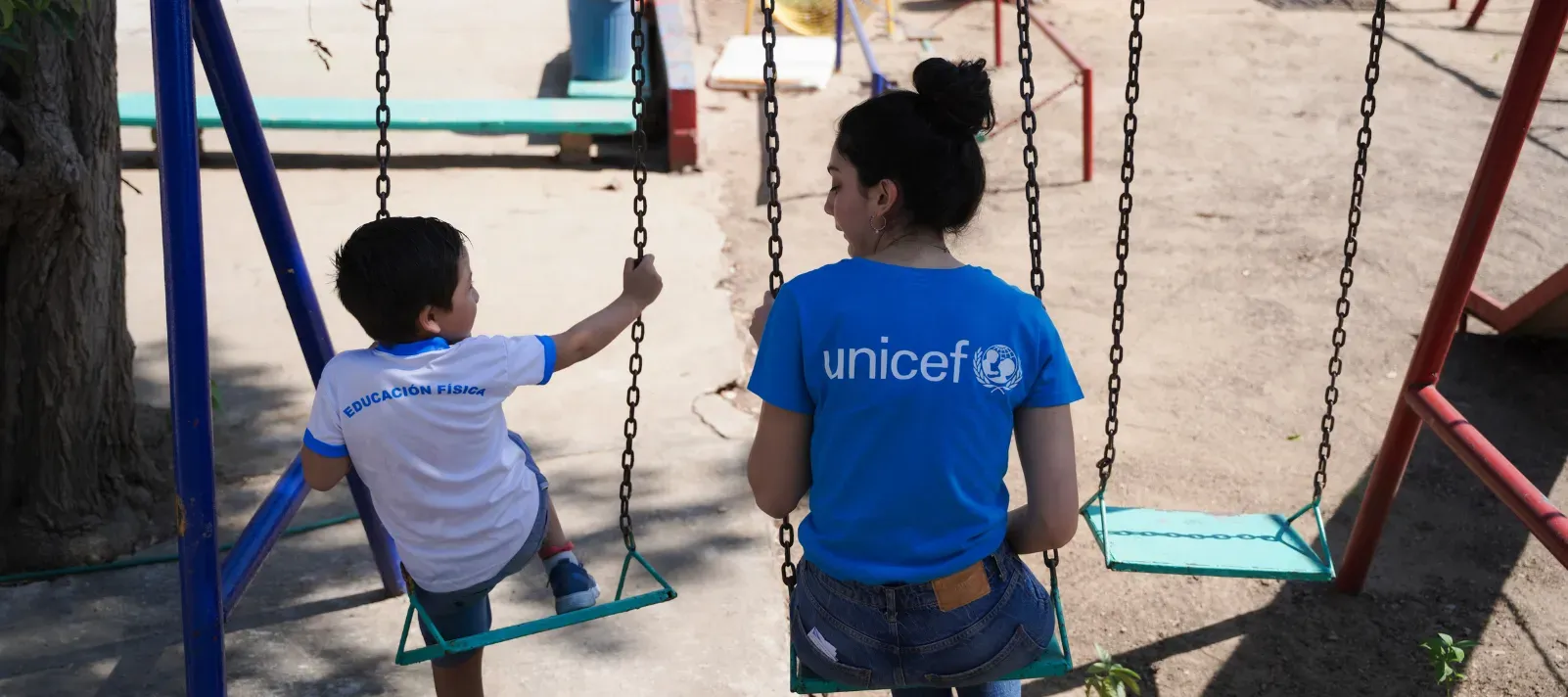 Trabajadora de UNICEF sentada en un columpio junto a un ni&ntilde;o peque&ntilde;o en el patio de juegos de una escuela, ambos de espaldas a la c&aacute;mara