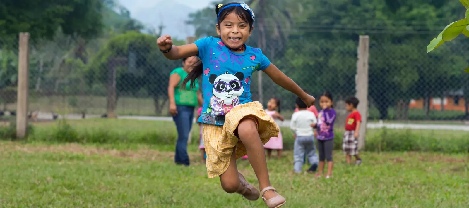 Una ni&ntilde;a con una camiseta azul est&aacute; saltando en una cancha en el exterior, detr&aacute;s de ella est&aacute;n otros ni&ntilde;os jugando