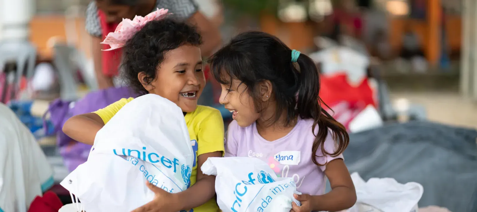  Dos ni&ntilde;as peque&ntilde;as sonriendo y sosteni&eacute;ndose mutuamente mientras cargan bolsas con el logo de UNICEF y art&iacute;culos de higiene