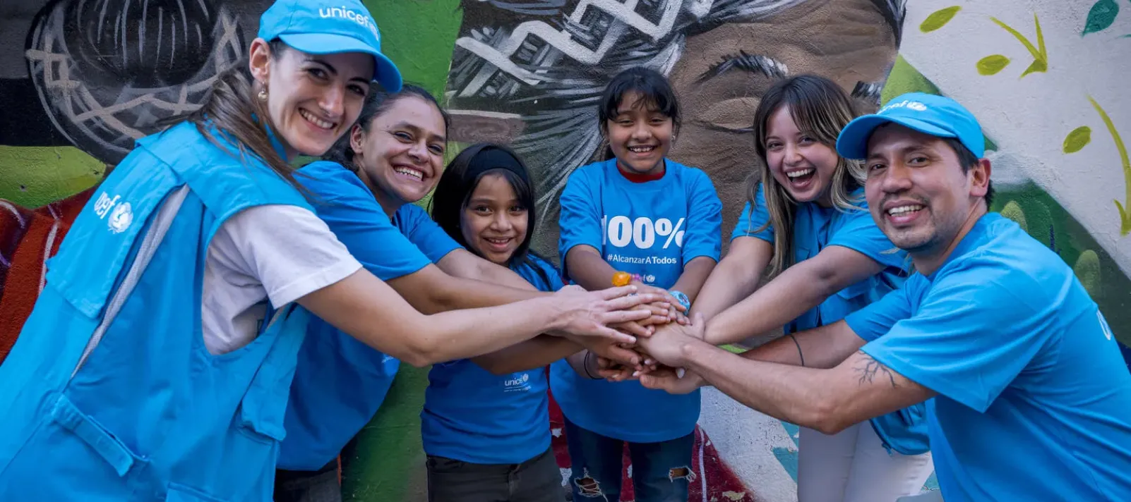 Funcionarios de UNICEF M&eacute;xico y ni&ntilde;as juntan sus manos frente a un mural, en se&ntilde;al de trabajo conjunto por la infancia.