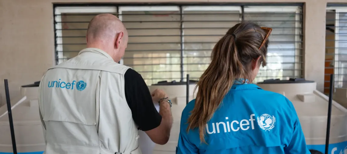 Dos integrantes del equipo de UNICEF, con chaleco y camisa institucionales, revisan informaci&oacute;n en una instalaci&oacute;n.