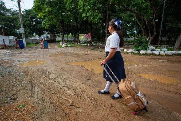Una ni&ntilde;a con su mochila en medio de un camino enlodado