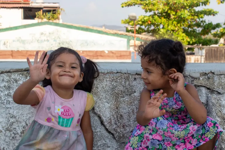 Dos ni&ntilde;as peque&ntilde;as sonr&iacute;en y saludan con la mano sentadas junto a una barda de piedra en un entorno urbano. La ni&ntilde;a de la izquierda, con vestido rosa, sonr&iacute;e ampliamente a la c&aacute;mara mientras saluda con la mano; la de la derecha, con vestido floral, la mira sonriendo.