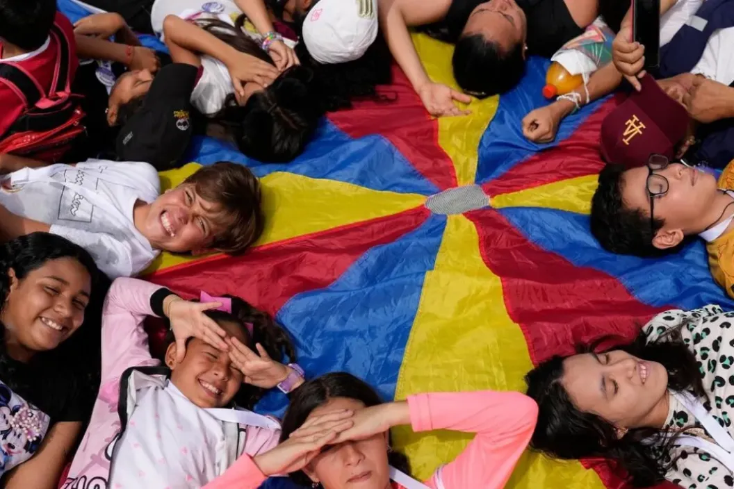Grupo de ni&ntilde;as, ni&ntilde;os, adolescentes y personas adultas recostados en c&iacute;rculo sobre una tela de paraca&iacute;das de colores, sonriendo y jugando juntos al aire libre.