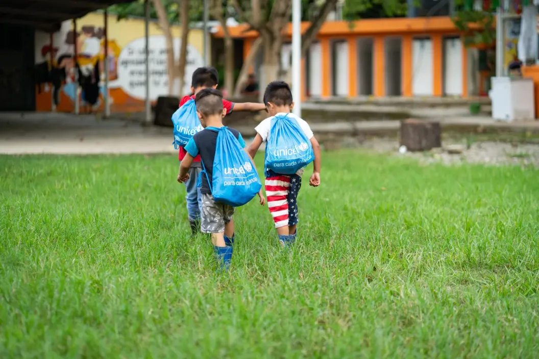 Tres ni&ntilde;os caminan por un prado verde y est&aacute;n de espaldas con tulas azules que tienen el logo de UNICEF 