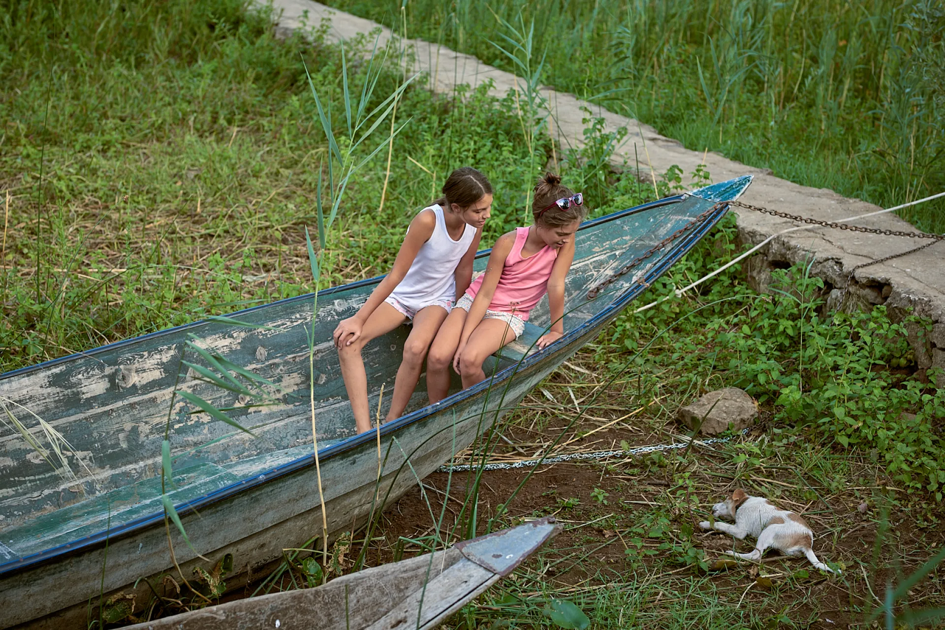 Two girls sitting in a boat on the ground watching a lying dog