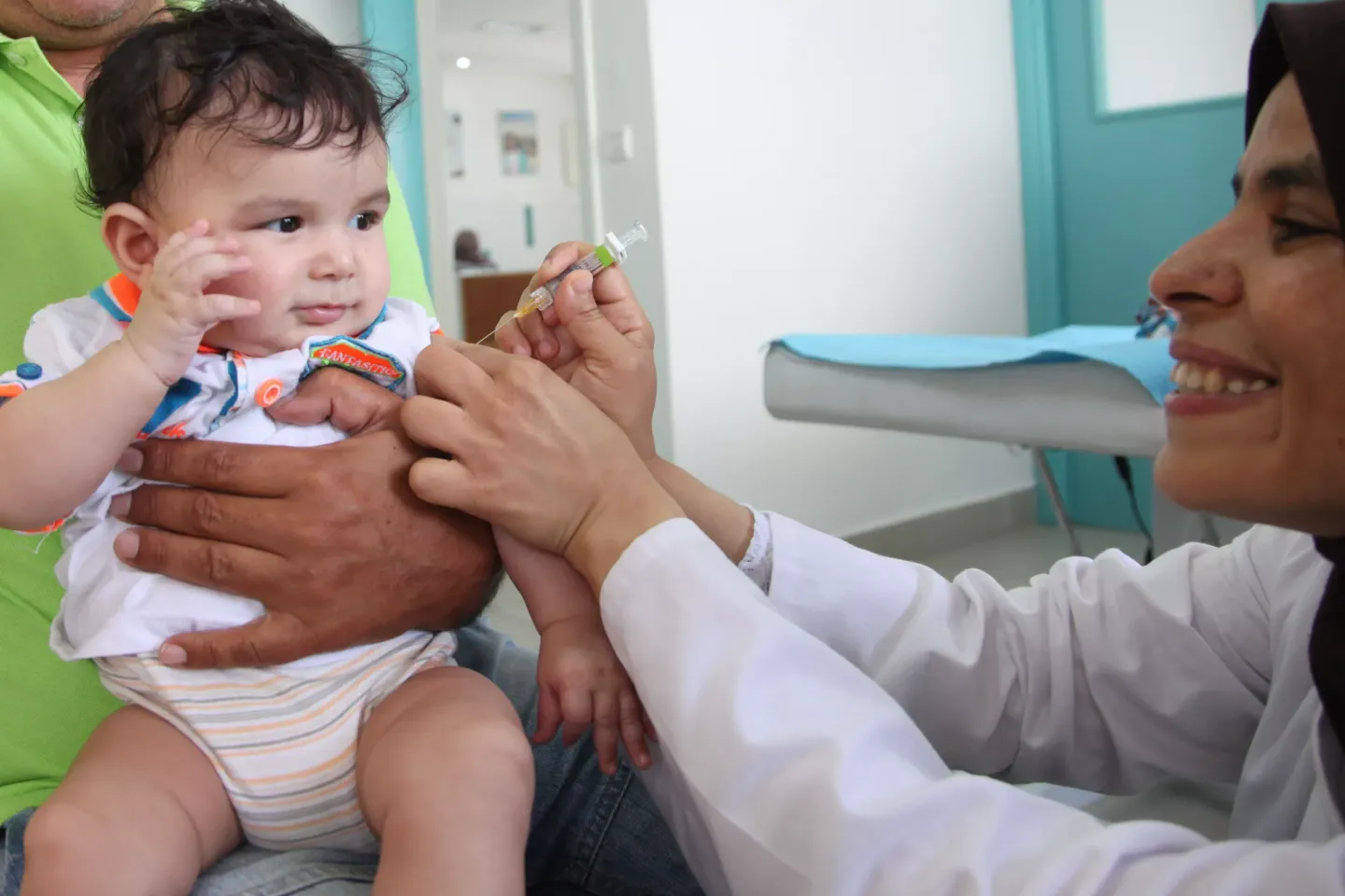 Une infirmi&eacute;re souriante, portant une blouse blanche, vaccine un jeune enfant dans une clinique m&eacute;dicale. L&rsquo;enfant semble l&eacute;g&egrave;rement surpris tandis que son p&egrave;re, v&ecirc;tu d&rsquo;une chemise verte, le tient. La sc&egrave;ne se d&eacute;roule dans un environnement m&eacute;dical moderne