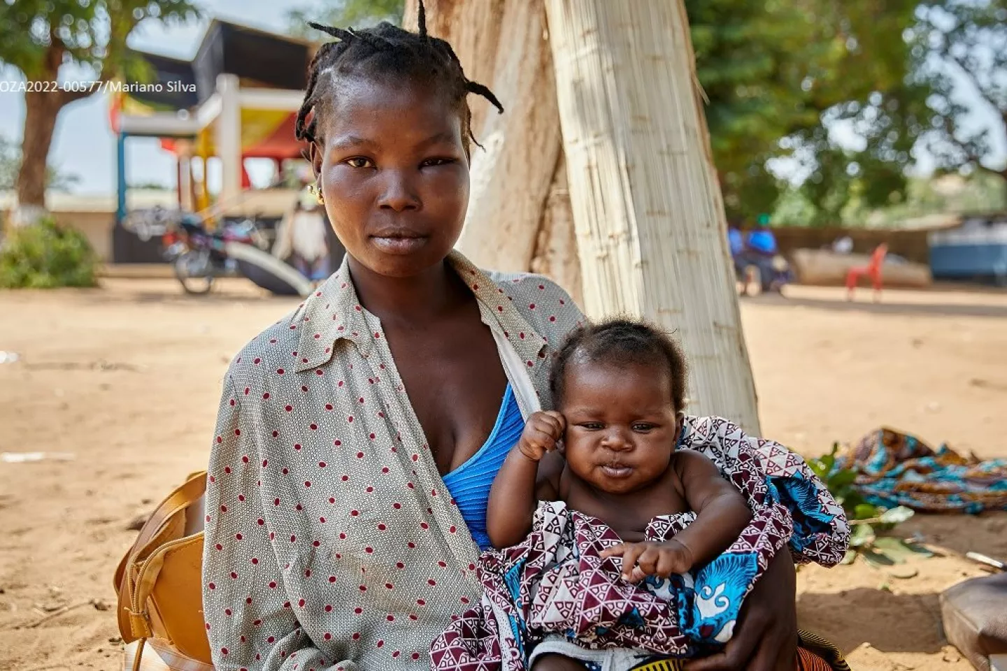 A young mother, with a swelling in her left eye and cheek, seats on the floor with her baby girl on her lap.