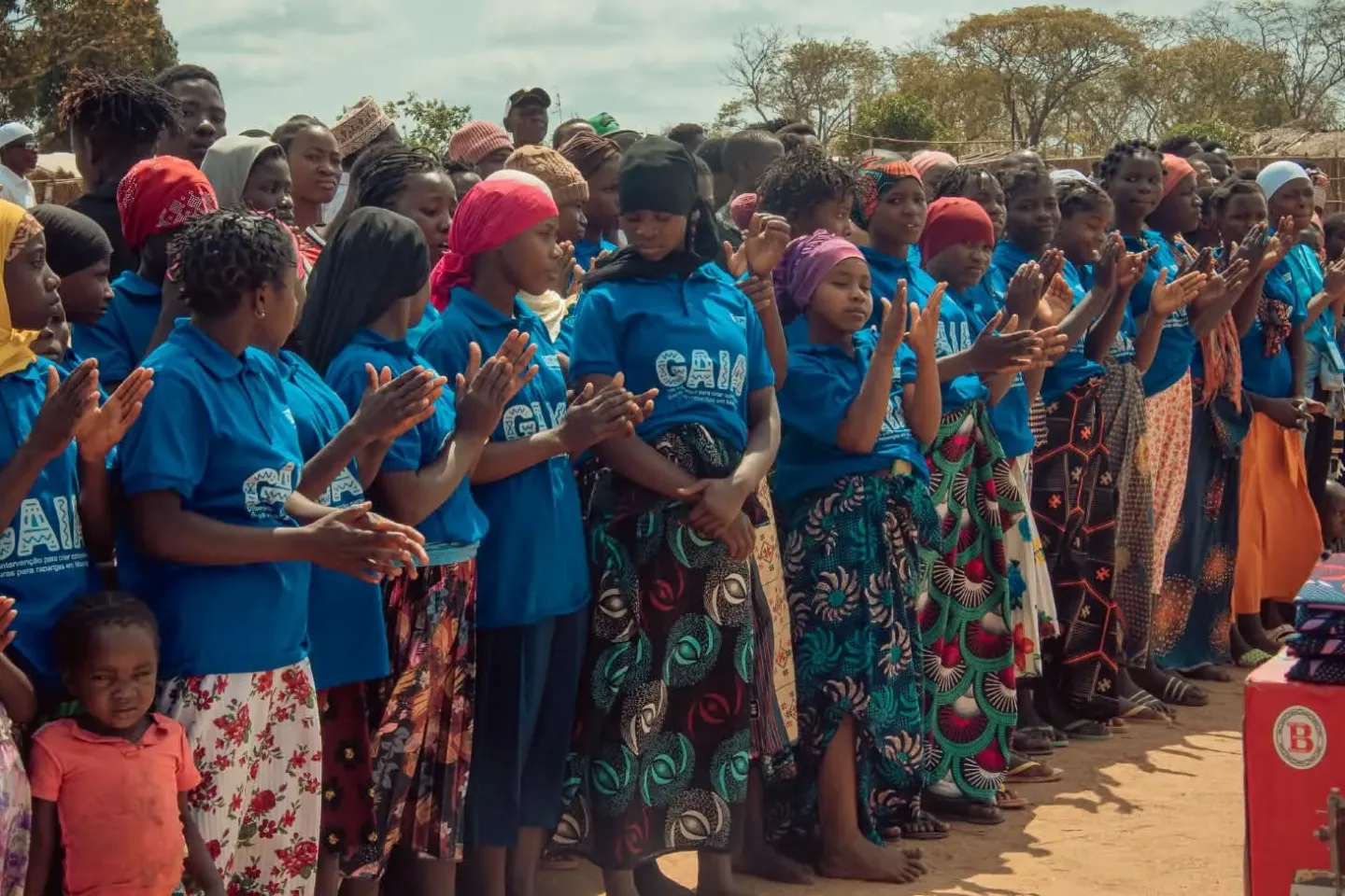 A group of women and girls wearing blue shirts and colorful skirts standing in a line outdoors, clapping and engaging in a community event.