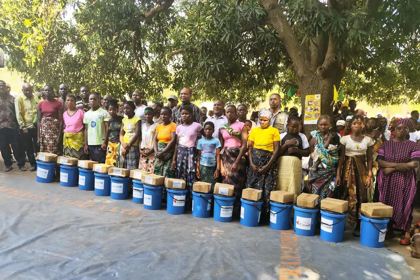 Group of men, women, and children standing behind blue buckets with supplies under a large tree outdoors.