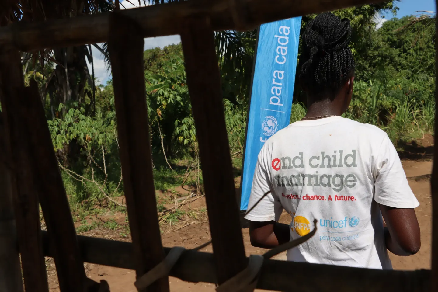 Uma menina vestida com uma camisa branca com a mensagem &laquo;end child marriage&raquo;, em p&eacute; ao ar livre junto a um cartaz da UNICEF, num cen&aacute;rio rural.