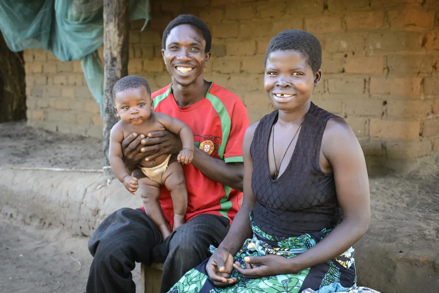 Uma fam&iacute;lia de tr&ecirc;s pessoas, sorridente, sentada &agrave; porta de uma casa r&uacute;stica de tijolo; o pai segura um beb&eacute; e a m&atilde;e est&aacute; sentada ao lado deles.